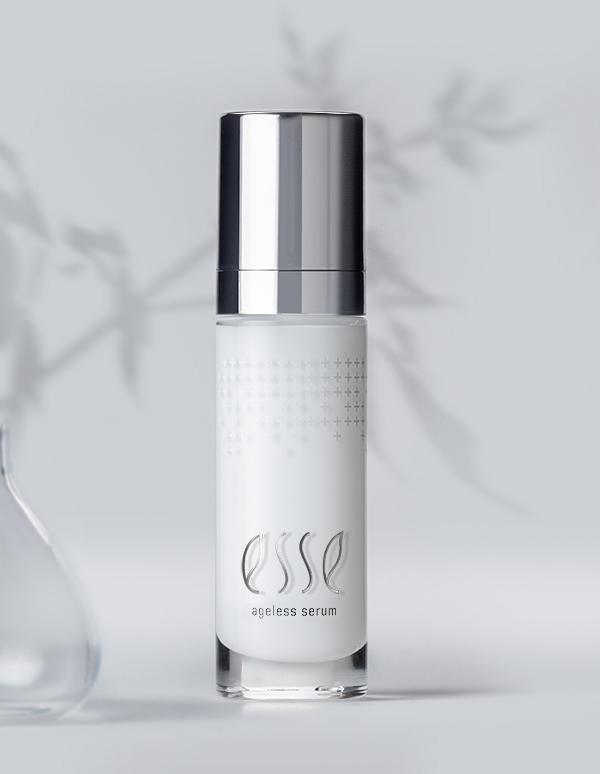A sleek, white bottle of Esse Ageless Serum with a metallic silver cap is showcased against a soft, white backdrop adorned with blurred botanical patterns. The bottle prominently features the brand name "Esse" and the phrase "Ageless Serum" in a subtle, elegant font.