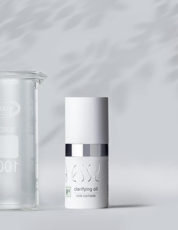 A white bottle of Esse Clarifying Serum is displayed on a light gray surface. The bottle has a silver band near its middle, and the text "P3 clarifying oil" is visible. A glass beaker is placed to the left of the bottle, partially obscured.