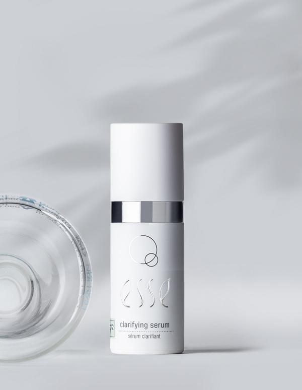 A white bottle labeled with "Esse Clarifying Serum" stands upright on a light gray surface. The bottle exhibits a sleek, minimalist design, accented by a silver band and distinctive black and green text. A shadow and part of a glass bottle are visible on the left.