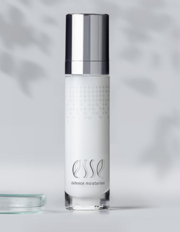 A sleek, cylindrical bottle of Esse Defence Moisturiser showcases a silver cap and minimalist design. The bottle contains white lotion and prominently displays the brand name "esse" along with the product name "defence moisturiser" on its front. The subtly blurred background enhances its elegant appearance.