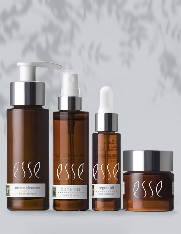 Four brown glass skincare bottles with white labels, part of the "Esse Dry Skin Trial/Travel Set," are arranged in a row. The products are, from left to right: cream cleanser, biome mist, repair oil, and a jar that might contain a cream or balm. The background is light grey with a subtle leafy pattern.