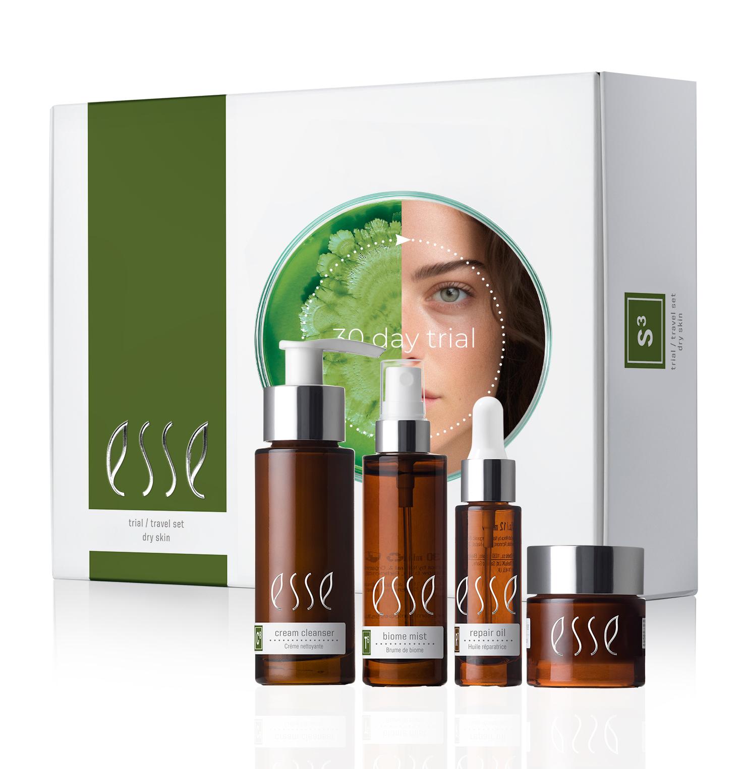 An image of the "Esse Dry Skin Trial / Travel Set" skincare collection. The set includes four brown bottles of various sizes and shapes labeled "cream cleanser," "biome mist," "repair oil," and "rich moisturizer." The packaging features a partial face and green plant.