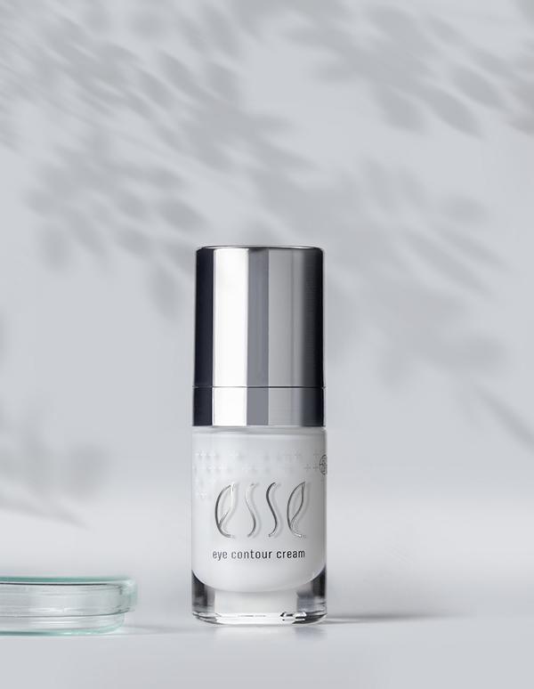A small bottle of Esse Eye Contour Cream with a sleek silver cap is placed against a minimalist background with dappled shadow patterns. A glass object is partially visible in the lower left corner.