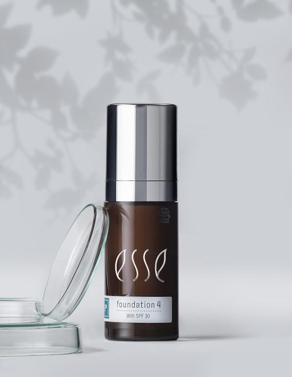 A bottle of Esse Foundation with SPF 30 stands on a clean, light gray surface. The dark bottle features a silver cap and displays its product details on the label. Beside it, a clear, round container is partially open. In the background, soft shadows of leaves create an elegant ambiance.