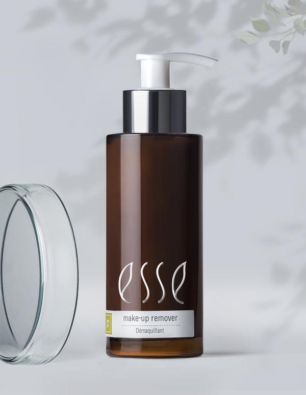 A brown bottle of Esse Make-up Remover with a white pump and a gray label stands against a light background with delicate shadowed leaves. The bottle's cap is also visible near the bottom left, and the product label includes the word "Démaquillant.