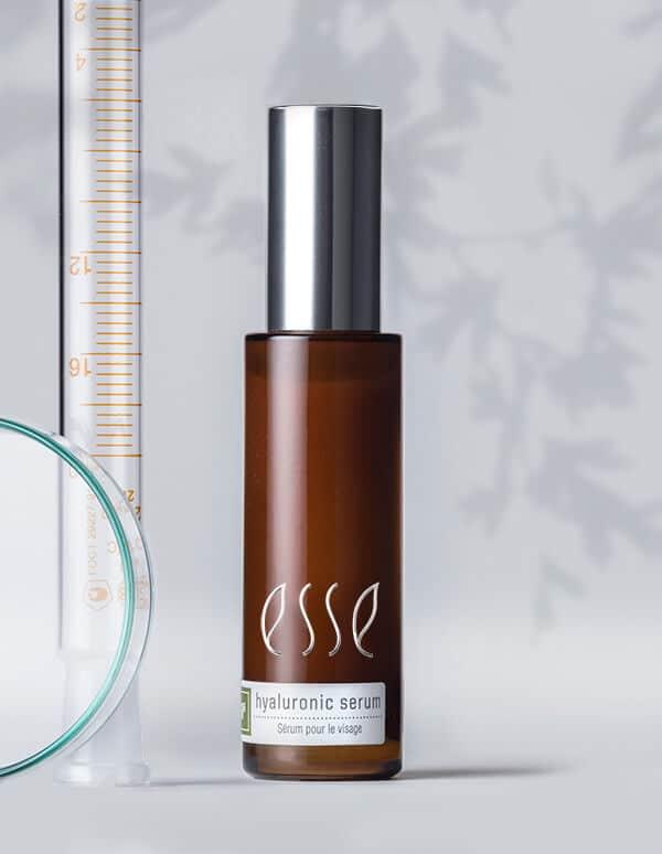 A brown bottle with a silver cap, labeled "Esse Hyaluronic Serum" and featuring the words "Serum pour le visage" underneath. The bottle is placed next to a glass petri dish and a vertical syringe, set against a light background with delicate leaf shadows.