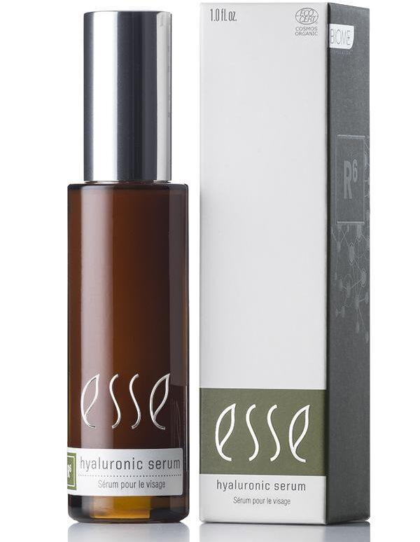 An Esse Hyaluronic Serum, encased in a brown bottle with a silver cap and labeled with white text, is displayed alongside its packaging. The box is predominantly white with one dark gray side, showcasing the same white text as on the bottle plus some extra details.