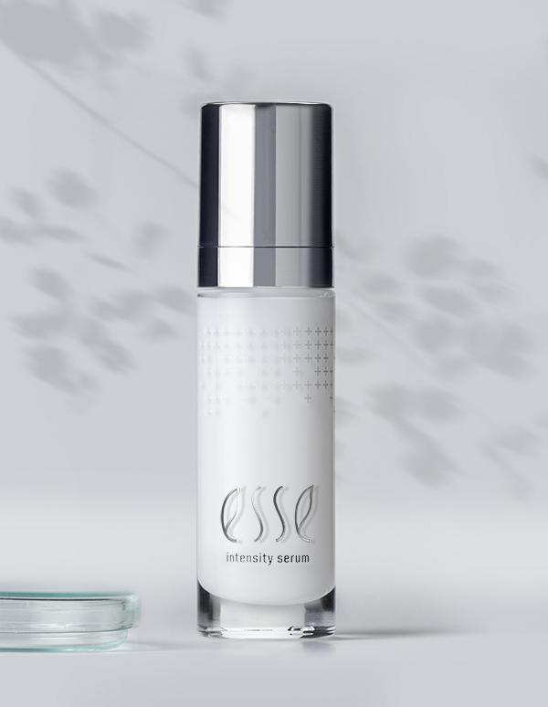 A bottle of Esse Intensity Serum, featuring a sleek white finish and a silver cap, stands upright against a light gray background. Subtle, blurred leaf shadows in the background enhance the natural aesthetic of the minimalist scene.