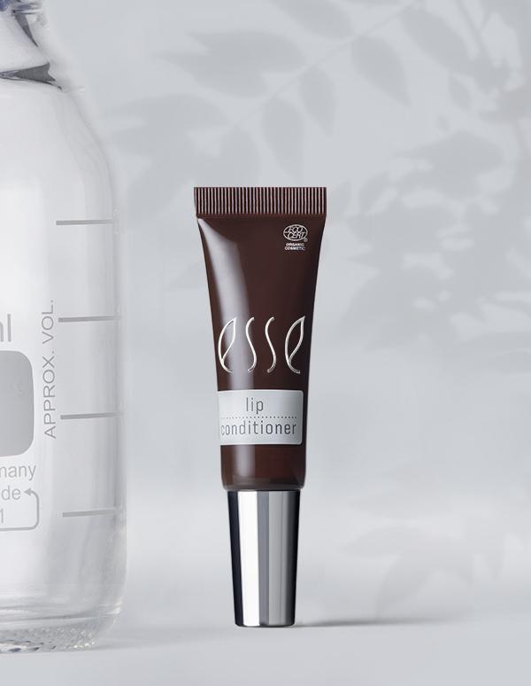 A brown tube of Esse Lip Conditioner with a silver cap stands upright. Positioned in front of it is a translucent container marked with measurements on its left side. The background features a softly lit setting accentuated by leaf shadows.