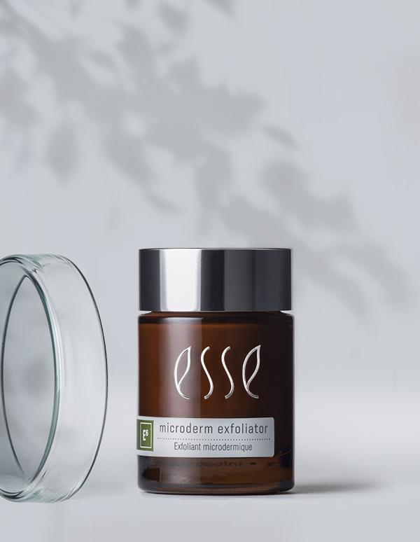 A jar of Esse Microderm Exfoliator with a brown body and a silver lid is positioned next to an open clear glass lid. The product label features text in both English and French, set against a light, blurred background adorned with leaf shadows that create a gentle, natural atmosphere.