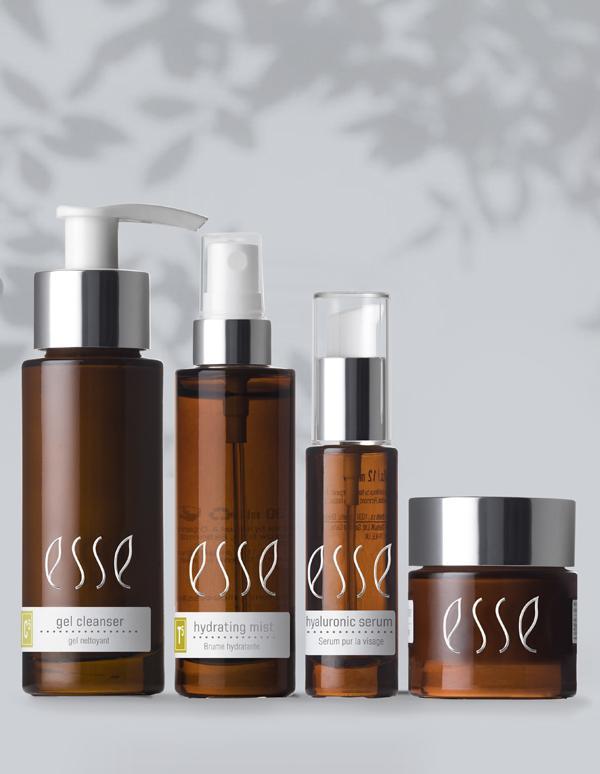 A row of four brown glass skincare bottles with white labels from the "Esse Oily / Combination / Normal Trial / Travel Set" is neatly arranged. From left to right: a gel cleanser with a pump, a hydrating mist with a sprayer, a hyaluronic serum with a smaller pump, and a cream in a jar. A shadow of foliage is cast above.