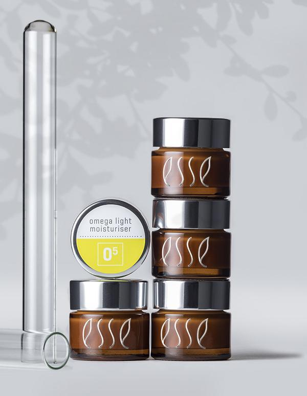 The image shows four brown "Esse" jars with silver lids stacked on top of each other. Next to them is a smaller jar labeled "Esse Omega Light Moisturiser." A clear glass test tube lies horizontally in front of the jars.