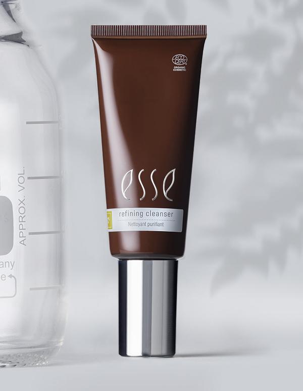 A tube labeled "Esse Refining Cleanser" with a silver screw cap in elegant brown is displayed against a minimalistic background. The product features text suggesting it is eco-friendly or organic. A blurred partial view of a clear bottle is visible to the left.