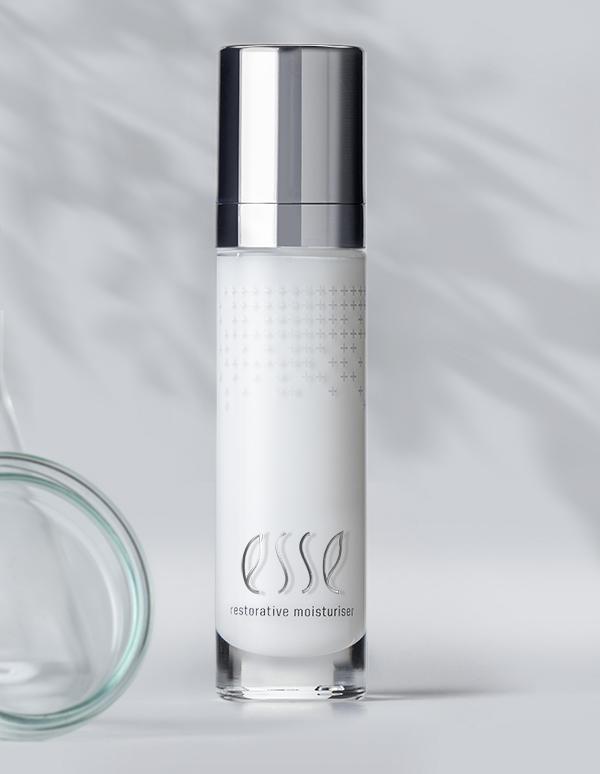A sleek, silver-capped bottle of Esse Restorative Moisturiser stands against a light grey background. The bottle boasts a minimalist design with subtle text and a faint dot pattern. Shadows from leaves are cast on the background, adding a natural touch.