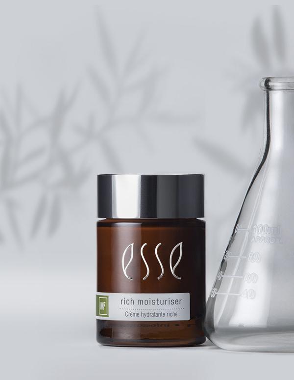 A glass jar of Esse Rich Moisturiser, featuring a brown-tinted body and silver cap, is positioned beside a laboratory glass beaker. The background is light gray with the faint shadow of foliage, creating a natural, scientific aesthetic.