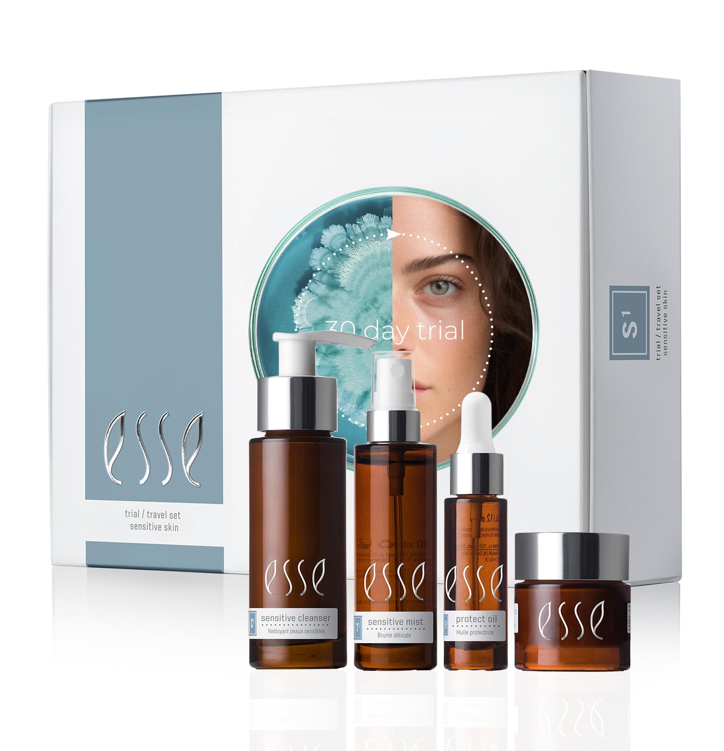 The Esse Sensitive Skin Trial / Travel Set is displayed, featuring four brown bottles and jars in various sizes positioned in front of white and blue packaging. The packaging highlights an image of a woman’s face partially obscured by a flower.