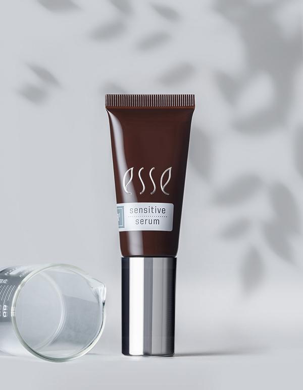 A tube of Esse Sensitive Serum stands upright against a light gray background with dappled shadows resembling leaves. The elegantly designed brown and silver packaging is paired with a clear, empty glass beaker resting on its side to the left.