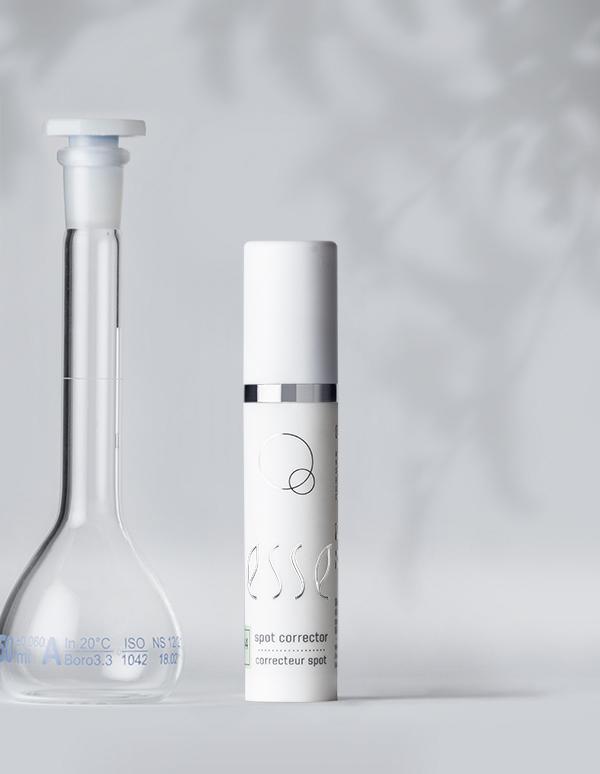 A clear lab flask with a white stopper stands beside a sleek white cylindrical bottle labeled "Esse Spot Corrector" on a light gray background. The minimalist design emphasizes clarity and purity, suggesting a focus on skincare or cosmetic products.