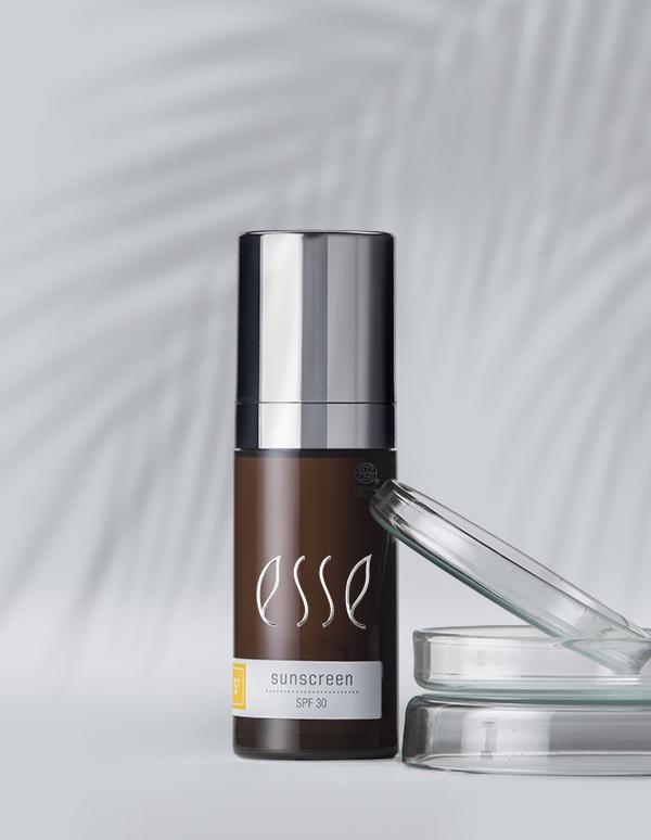 An Esse Sunscreen bottle with SPF 30 is showcased against a light background, accented by the shadow of a palm leaf. The dark-colored bottle features silver accents, and an open clear lid rests beside it.