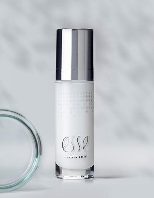A sleek bottle of Esse probiotic serum with a metallic pump dispenser stands upright. The branding text "Esse probiotic serum" is visible on the frosted glass body. The cap and background are shades of gray and white, giving the image a clean, minimalistic look.