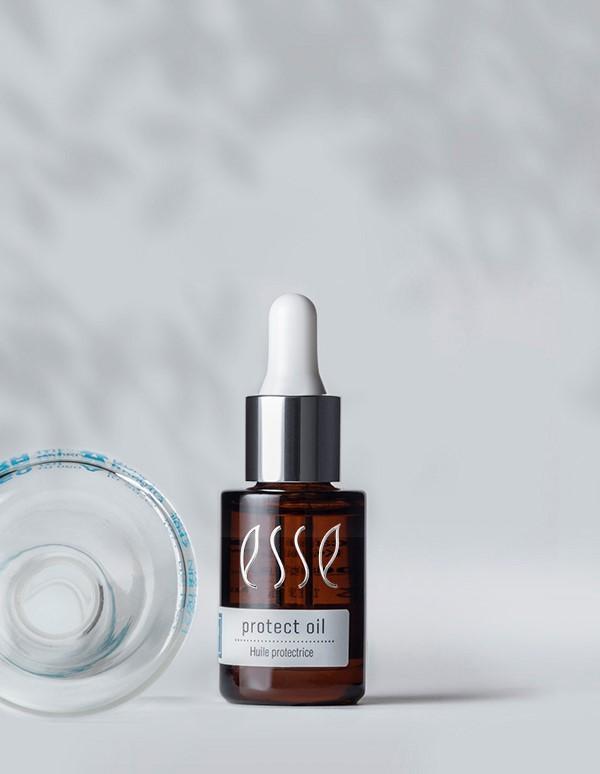 A brown bottle of "Esse Protect Oil" with a white dropper cap is standing next to a clear, open glass jar. The background is light and blurred with soft grey shadows.