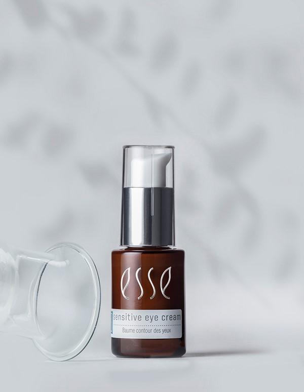 A small brown bottle of Esse Sensitive Eye Cream with a white pump dispenser and a clear cap is shown. The label on the bottle reads "Sensitive eye cream Baume contour des yeux." The background is blurred with a subtle shadow pattern.