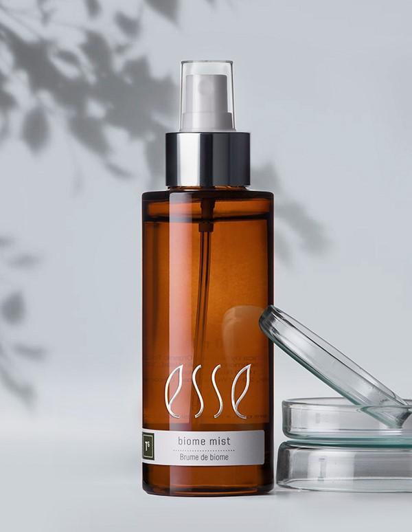 A brown glass bottle labeled "Esse Biome Mist" with a white and silver spray pump is resting on a light gray surface. A clear glass dish partially covers the bottle, while shadows of leaves in the background create a natural ambiance.