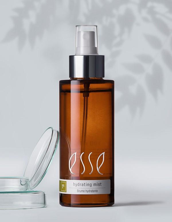 A bottle of Esse Hydrating Mist, featuring a white label and silver pump dispenser, stands next to a clear, open cap. The background is light grey with soft shadows of leaves in the top right corner. The brown bottle's reflection is faintly visible on the surface.