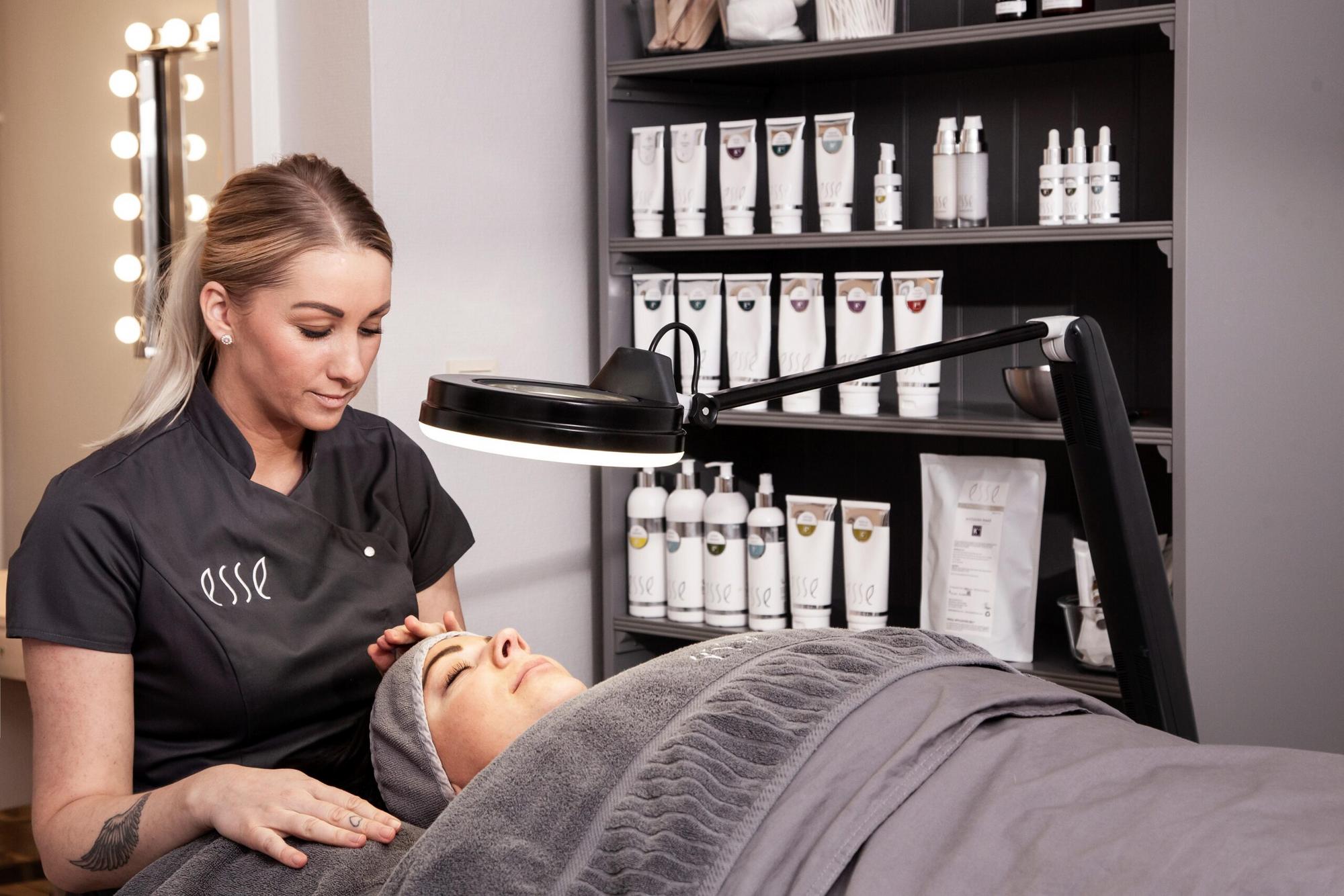 An esthetician wearing a gray uniform is giving a facial treatment to a client lying on a treatment table covered with gray blankets. The room has shelves stocked with various skincare products and a magnifying lamp positioned over the client's face.