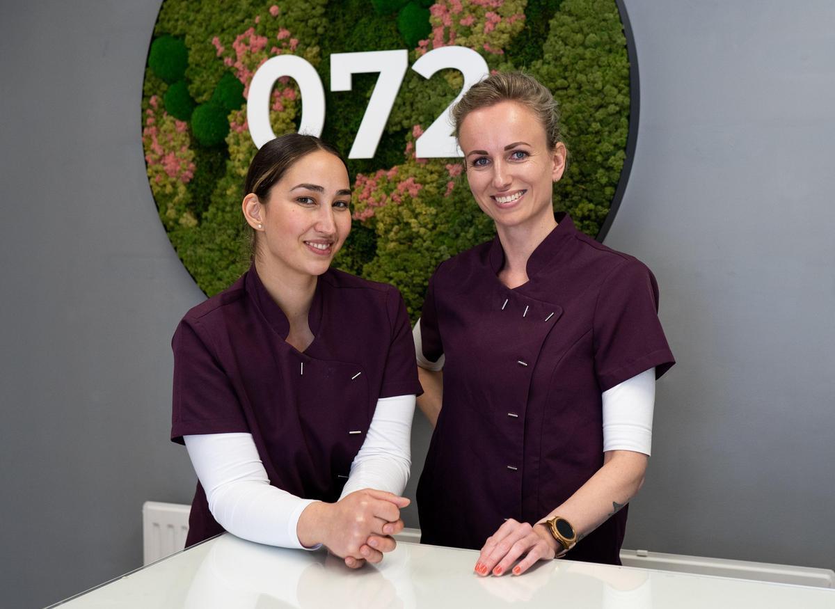 Two women in matching purple uniforms stand behind a white counter. One woman has light hair pulled back and the other has dark hair tied back. Both smile in front of a decorative wall display featuring the number "072" in white, surrounded by green and pink foliage.