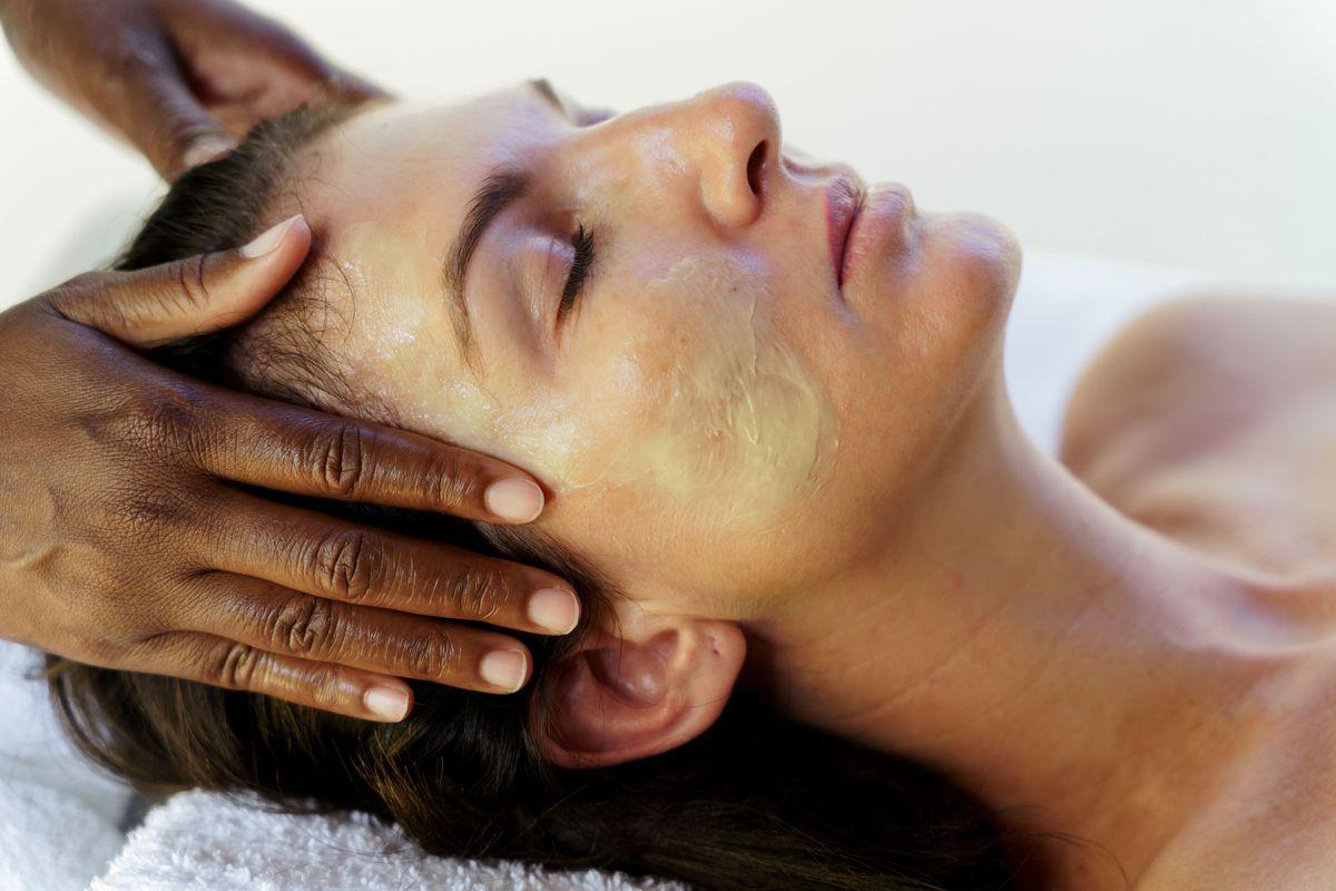 A person lying down with eyes closed is receiving a facial treatment. An individual with dark skin is gently massaging the person's face, which has a facial mask applied. The setting appears calm and relaxing.