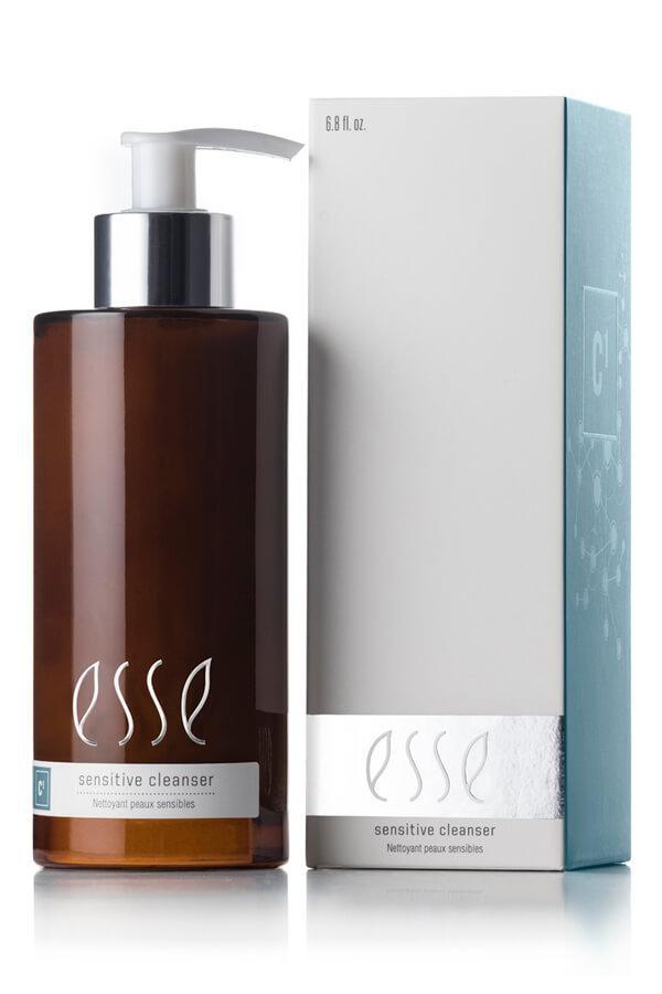 A bottle labeled "Esse Sensitive Cleanser" with a pump dispenser and brown in color is positioned next to a silver and white box that features the same branding. The bottle's label showcases white text, while the box has a metallic strip near the bottom providing product information.