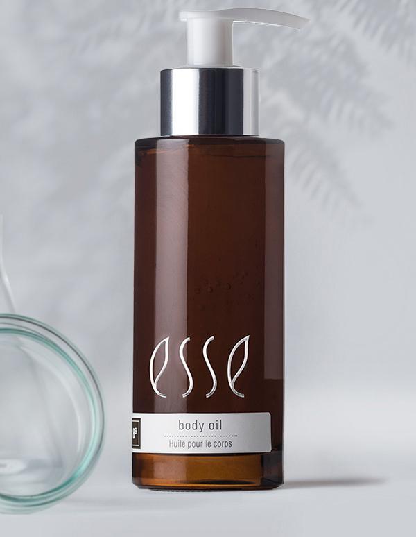 A brown bottle with a pump dispenser labeled "esse body oil - huile pour le corps." The bottle has sleek, minimalist design with a silver cap, set against a light background with a faint shadow of foliage.