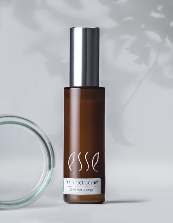 A brown cylindrical bottle labeled "resurrect serum" with a metallic silver cap stands upright on a light gray surface. The background is softly lit with blurred shadows, creating a minimalistic and elegant scene. An open lid is partially visible on the left.