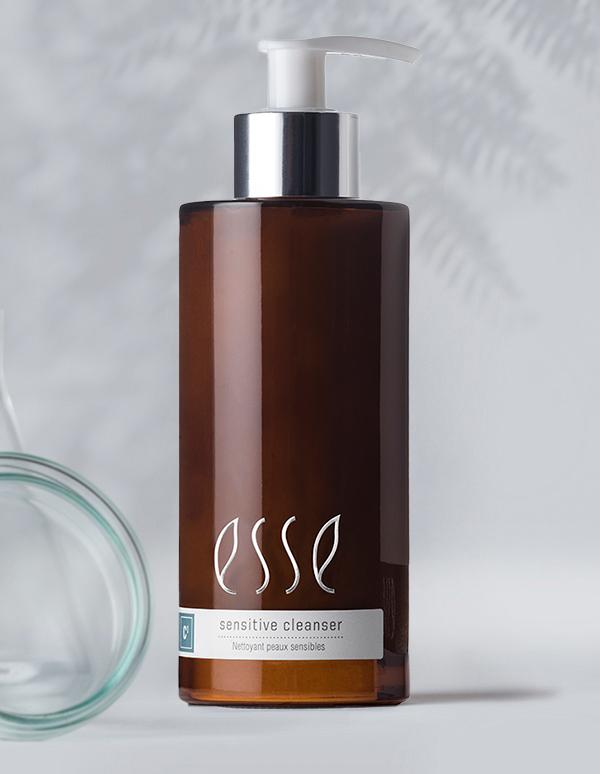 A brown bottle of Esse Sensitive Cleanser with a silver pump sits in front of a blurred background. The white label has the branding "Esse" and the text "Sensitive Cleanser - Nettoyant peaux sensibles." A glass object is partially visible on the left side.