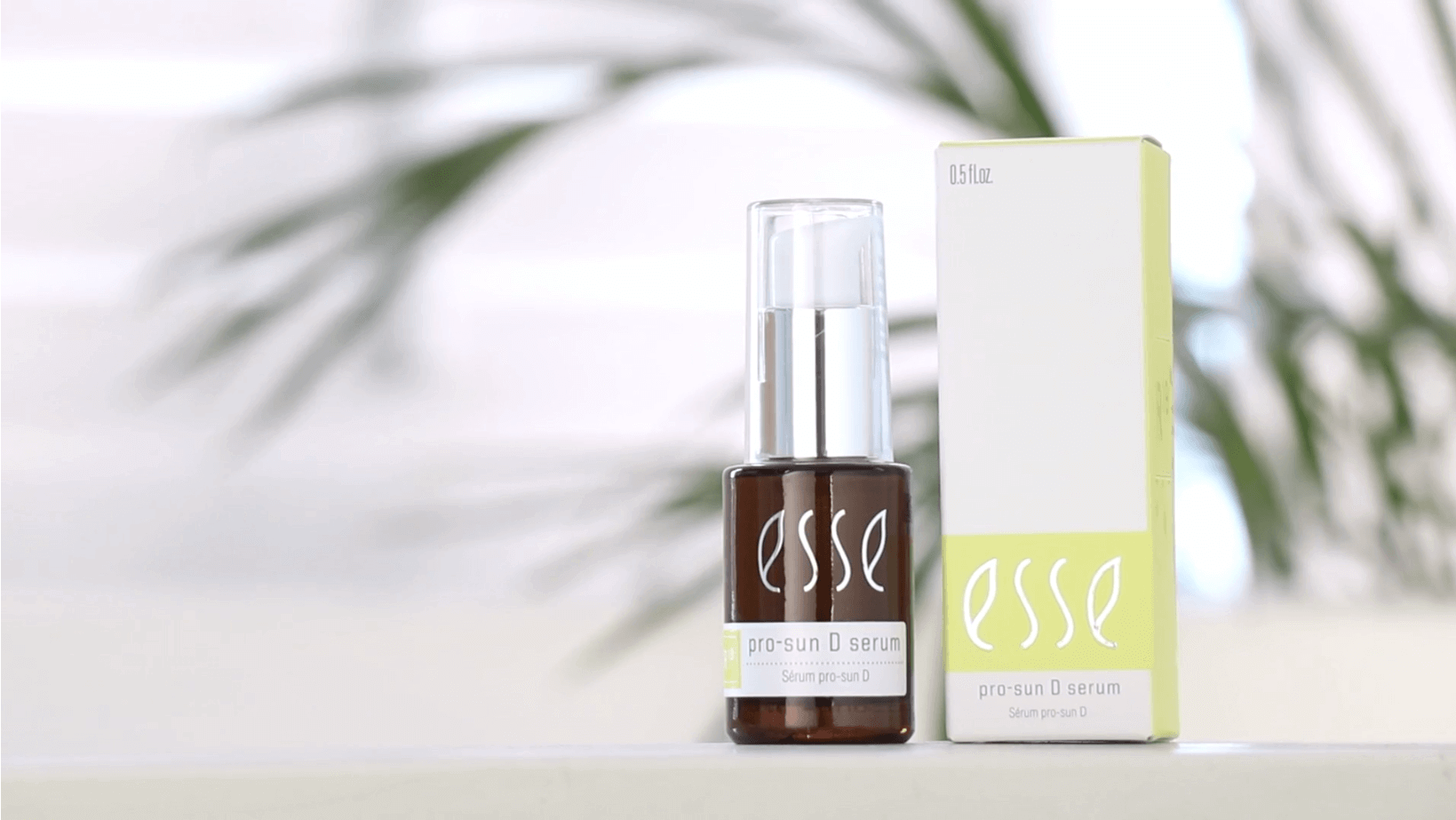 A compact, brown bottle of Esse Pro-Sun D Serum with white text on the label is shown alongside its corresponding green and yellow box. The serum bottle features a transparent cap. Both products are positioned on a white surface, set against a backdrop of blurred foliage.