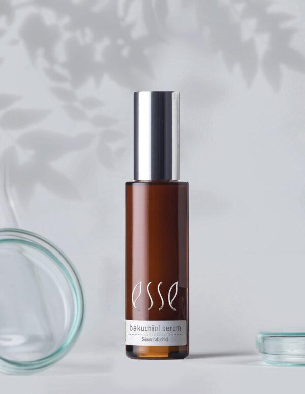 A brown glass bottle labeled "Esse Bakuchiol Serum" with a silver cap stands centered on a light grey surface. To the left, a lid enhances visual balance, while another lid lies flat in the bottom right corner. Shadows of branches add elegance to the background.