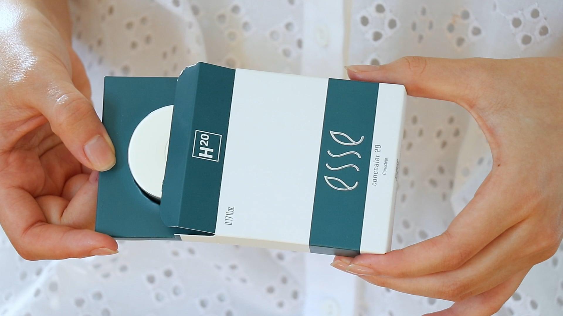 Close-up of a person holding a partially opened box of Esse Concealer skincare product. The white and teal packaging is shown against a background with a white, eyelet-textured fabric.