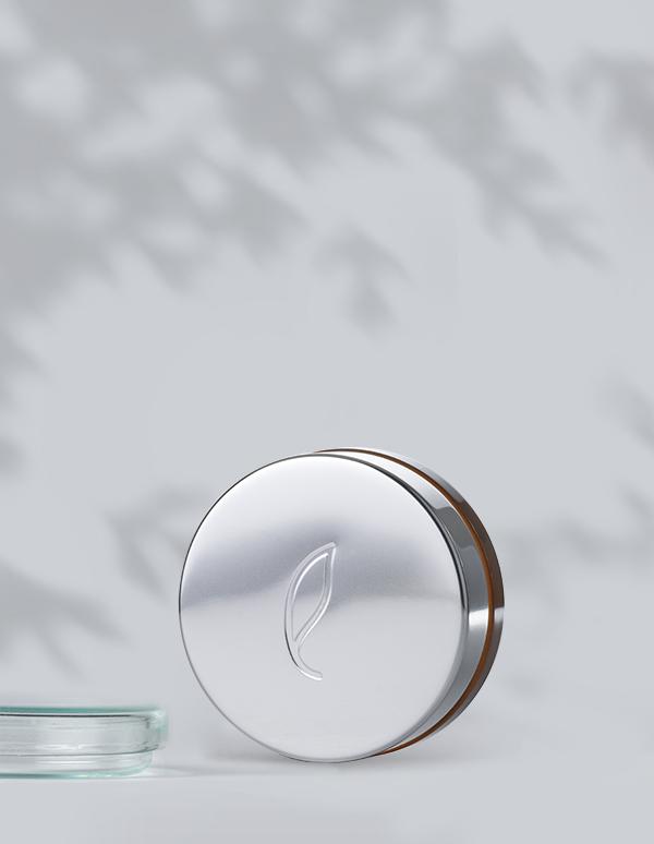 A round, silver tin labeled Esse Concealer with an embossed leaf design on the lid stands against a light gray background. The tin is partially open, revealing a hint of the concealer inside. Shadows of leaves create a subtle pattern on the wall in the background.