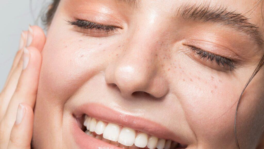 Close-up of a person with clear skin, freckles, and a bright smile. Their eyes are closed as they gently touch their face with one hand, showcasing radiant skin and natural beauty. The overall expression conveys happiness and satisfaction, emphasizing the effectiveness of the Esse Oily / Combination / Normal Trial / Travel Set.