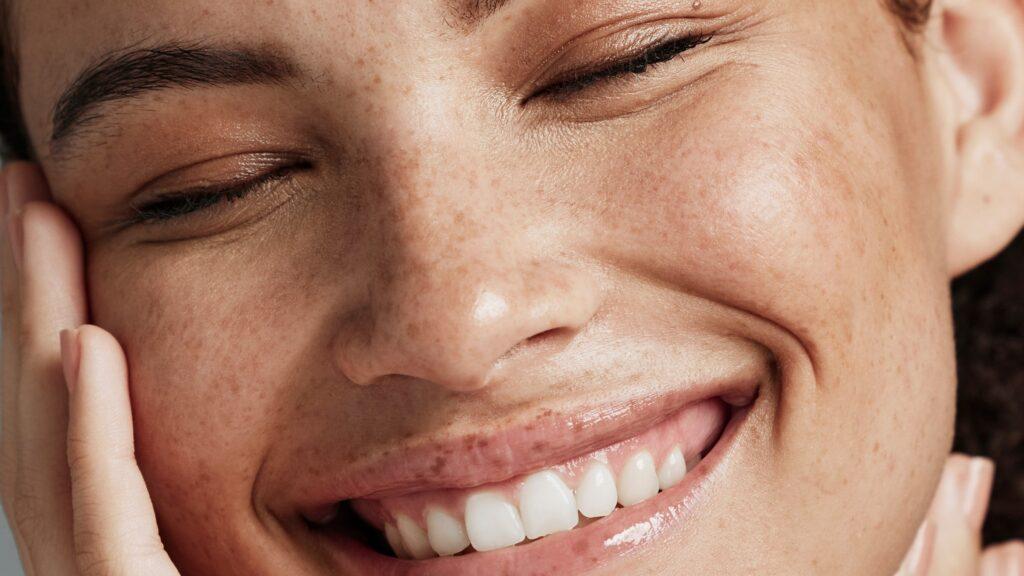 A close-up of a smiling person's face with eyes closed, showcasing the effects of the Esse Sensitive Skin Trial / Travel Set. The skin has visible freckles, and the teeth are showing in a wide smile. Their hand gently touches the side of their face, radiating happiness and contentment.