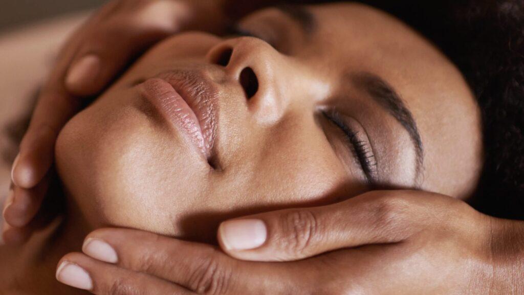 A close-up of a person with their eyes closed, appearing relaxed, as someone gently holds their face with both hands, possibly using the Esse Dry Skin Trial / Travel Set during a facial massage or spa treatment. The background is blurred, emphasizing a peaceful and soothing atmosphere.
