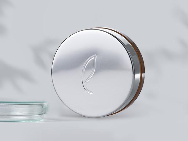 A round, metallic tin with a leaf symbol embossed on the lid sits on a light surface. The tin has a shiny appearance with a subtle reflection. To the left, a partial view of a clear glass object is visible. Shadows are cast on the background.