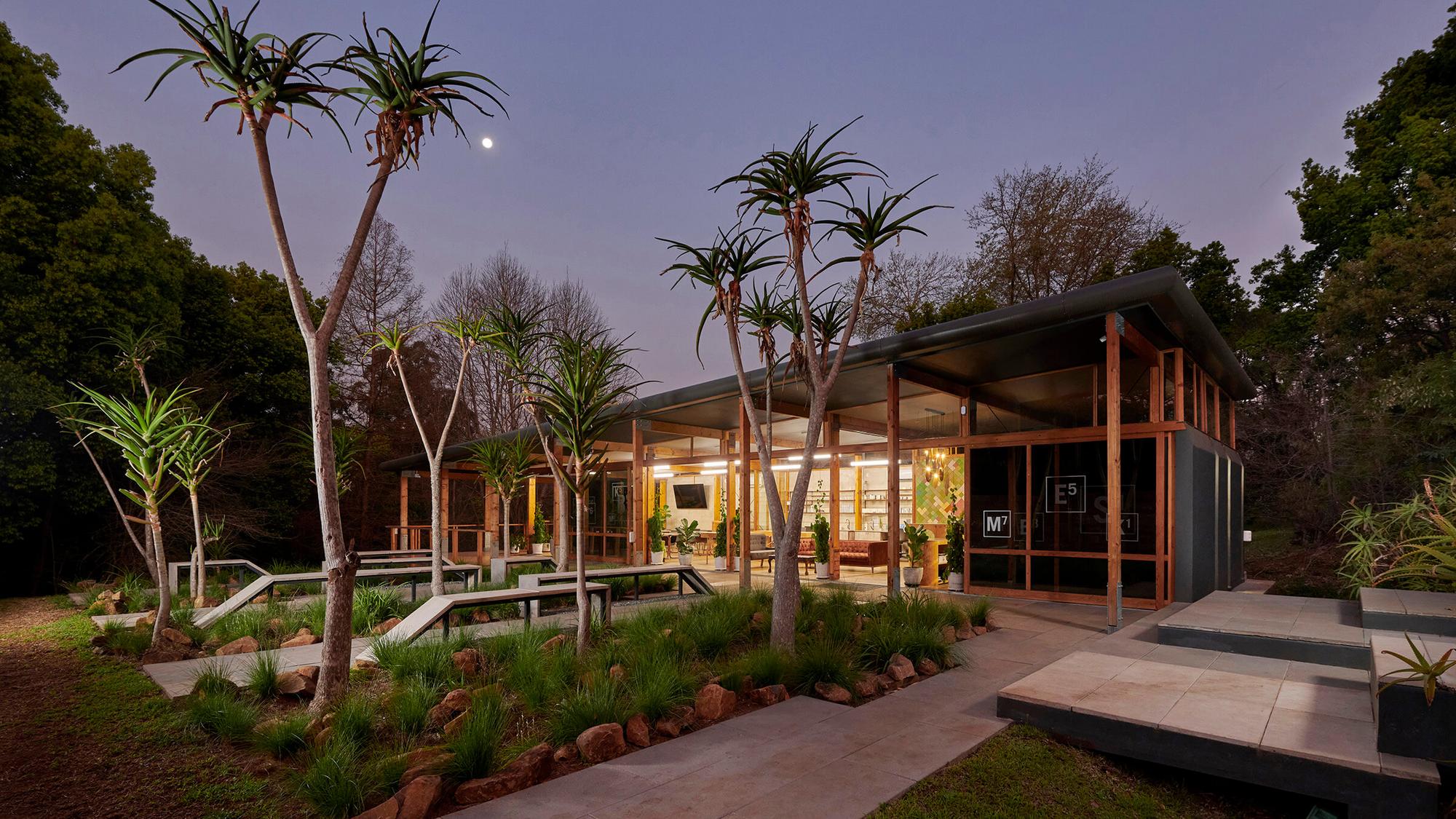 A modern glass building is surrounded by tall, spiky-leafed trees and lush greenery. The structure features large windows and geometric pathways, with warm interior lights visible. A crescent moon is visible in the dusk sky.