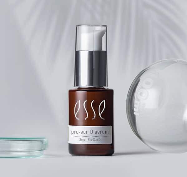 A brown bottle of Esse Pro-Sun D serum with a white pump sits on a light gray surface. The background features soft palm leaf shadows, and a clear round object is partially visible to the right.