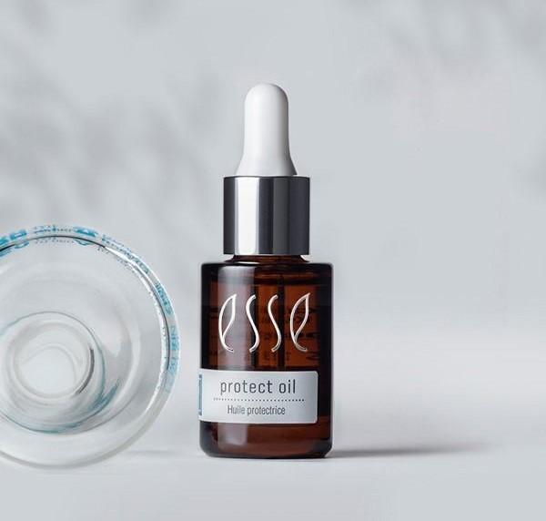 A brown dropper bottle labeled "Esse Protect Oil" stands upright with a silver cap and white dropper. Next to it, a clear, open lid lies on its side. The background is a soft gray with a subtle shadow pattern.